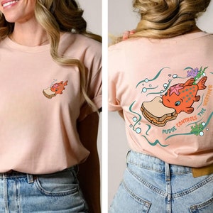 May include: A pink t-shirt with a cartoon illustration of a fish eating a sandwich. The text on the shirt reads "Pudge Controls the Weather".