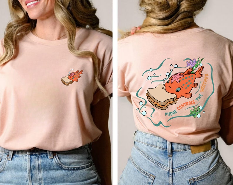 May include: A pink t-shirt with a cartoon illustration of a fish eating a sandwich. The text on the shirt reads "Pudge Controls the Weather".