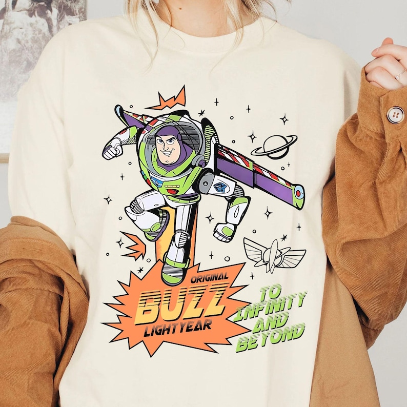 K&ouml;nnte beinhalten: Cremefarbenes T-Shirt mit einer Grafik von Buzz Lightyear in einer dynamischen Pose. Das Design enth&auml;lt den Text "Original Buzz Lightyear" und "To Infinity and Beyond" in Orange und Gr&uuml;n. Das Shirt hat einen Rundhalsausschnitt und lange &Auml;rmel.