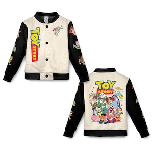 May include: Cream-coloured Toy Story bomber jacket with black sleeves and trim. The front features the Toy Story logo and character graphics. The back displays a group of Toy Story characters. The sleeves have character graphics.