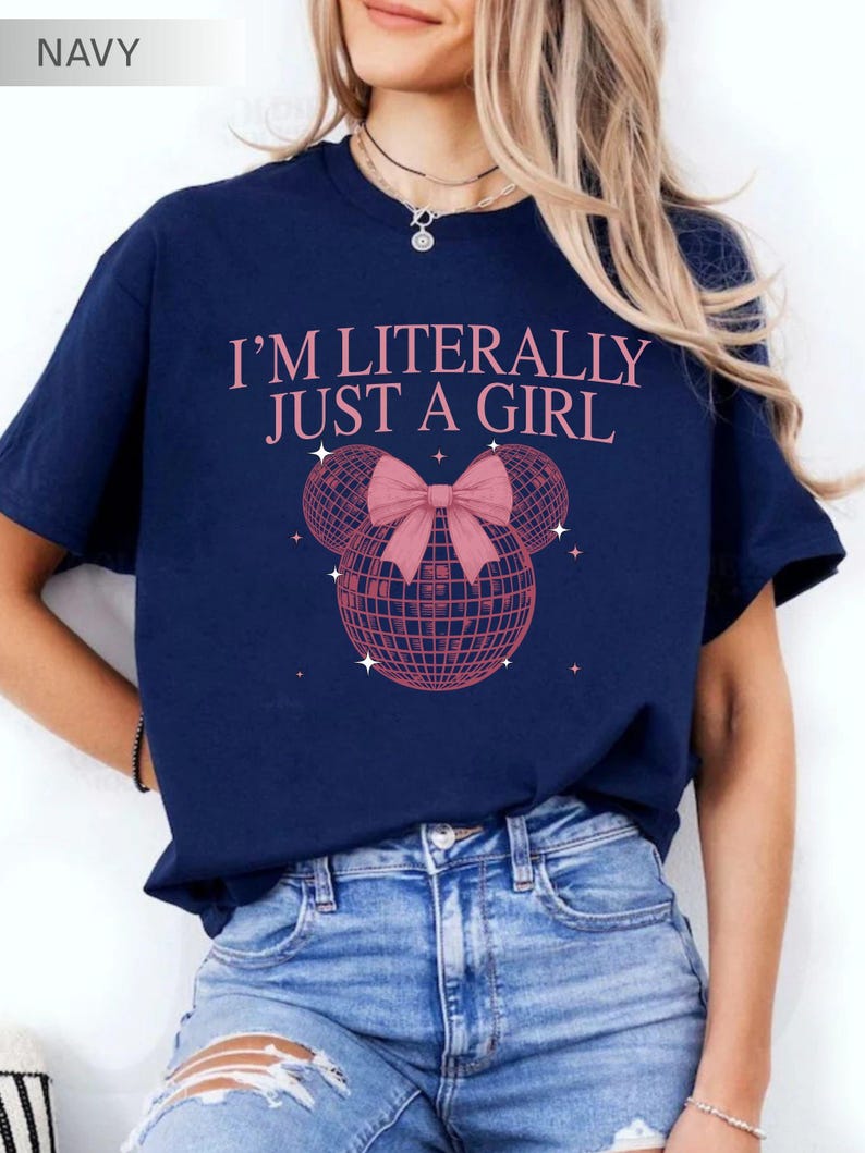 May include: Navy blue t-shirt with the text "I'M LITERALLY JUST A GIRL" in pink. The design includes a pink disco ball with a bow and star accents. The shirt is a casual style.