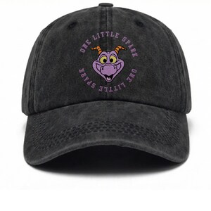 May include: A charcoal black baseball cap with a distressed finish. The front features an embroidered purple dragon face with orange horns, surrounded by the circular text "ONE LITTLE SPARK" in light purple lettering.