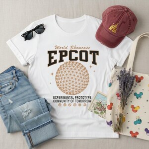 May include: White t-shirt with "EPCOT" graphic, a maroon baseball cap with a gold castle, and a canvas tote bag with colorful Mickey Mouse designs. The shirt also has the text "World Showcase" and "Experimental Prototype Community of Tomorrow".