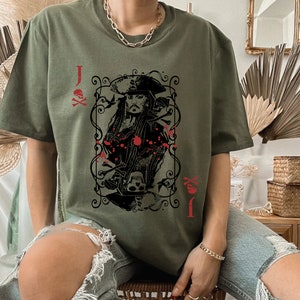 May include: Olive green t-shirt featuring a playing card design with a pirate theme. The card shows a portrait of a pirate captain, skulls, and crossbones, with the letter "J" and suit symbols in red.