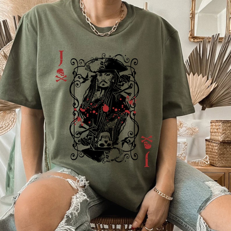 May include: Olive green t-shirt featuring a playing card design with a pirate theme. The card shows a portrait of a pirate captain, skulls, and crossbones, with the letter "J" and suit symbols in red.