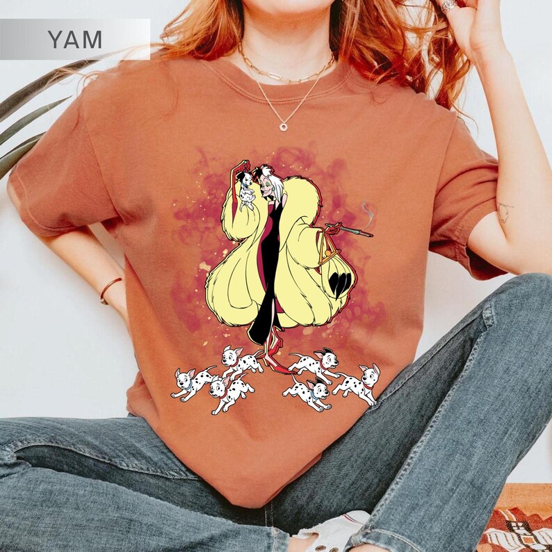 K&ouml;nnte beinhalten: Rostfarbenes Rundhals-T-Shirt mit einer Cartoon-Illustration von Cruella de Vil in einem gelben Mantel, die eine Zigarette h&auml;lt, mit Dalmatiner-Welpen. Das Wort "YAM" befindet sich in der oberen linken Ecke.