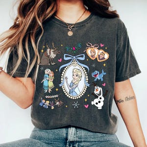 Puede incluir: Camiseta gris oscuro con un gr&aacute;fico colorido de personajes de una popular pel&iacute;cula animada. El dise&ntilde;o incluye a Elsa en un marco ovalado, un reno, Olaf el mu&ntilde;eco de nieve y otros personajes. La camiseta tiene cuello redondo.