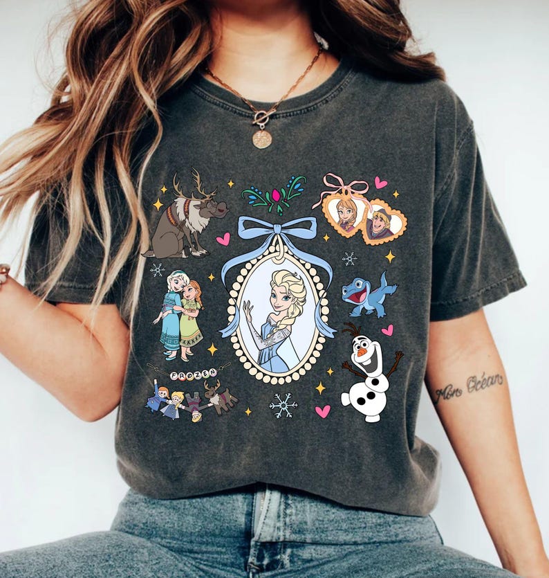 Puede incluir: Camiseta gris oscuro con un gr&aacute;fico colorido de personajes de una popular pel&iacute;cula animada. El dise&ntilde;o incluye a Elsa en un marco ovalado, un reno, Olaf el mu&ntilde;eco de nieve y otros personajes. La camiseta tiene cuello redondo.