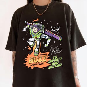 K&ouml;nnte beinhalten: Schwarzes T-Shirt mit einem farbenfrohen Buzz Lightyear-Motiv. Das Design enth&auml;lt die Worte "Original Buzz Lightyear" und "To Infinity and Beyond", sowie Sterne, einen Planeten und eine Rakete. L&auml;ssiger Stil.