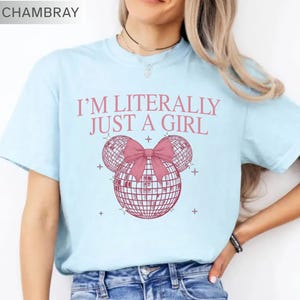 May include: Light blue t-shirt with the text "I'M LITERALLY JUST A GIRL" in pink. The shirt features a pink bow and two disco ball shapes. The word "CHAMBRAY" is in the top left corner.