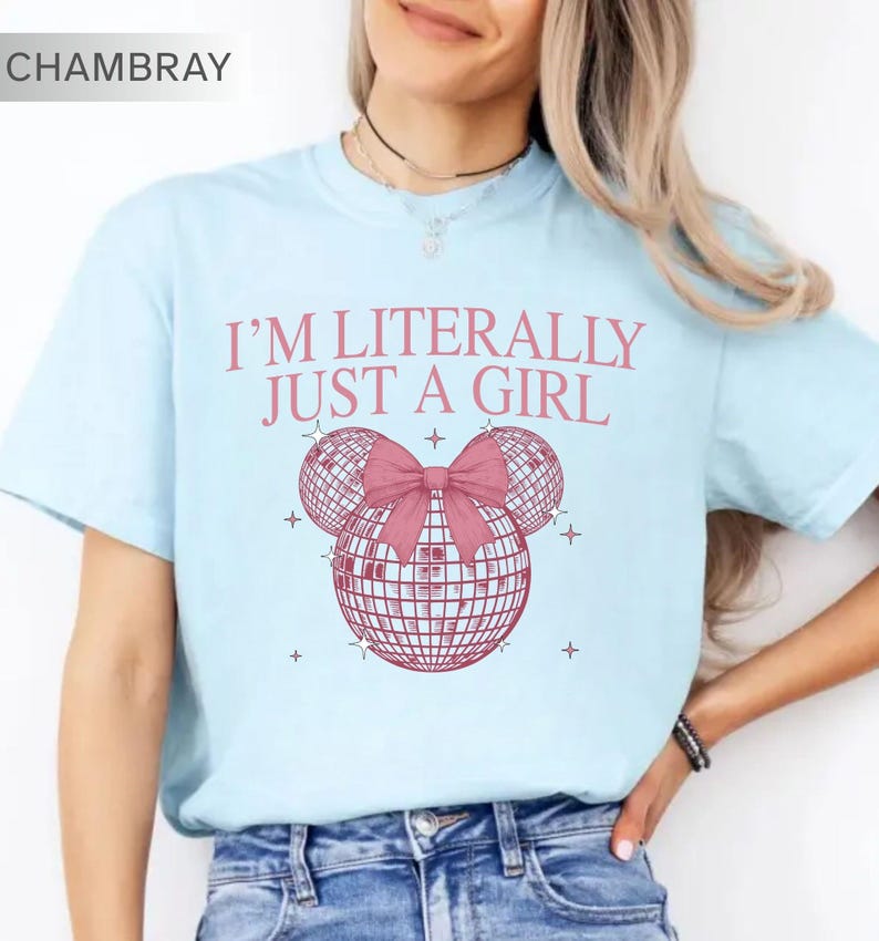 May include: Light blue t-shirt with the text "I'M LITERALLY JUST A GIRL" in pink. The shirt features a pink bow and two disco ball shapes. The word "CHAMBRAY" is in the top left corner.