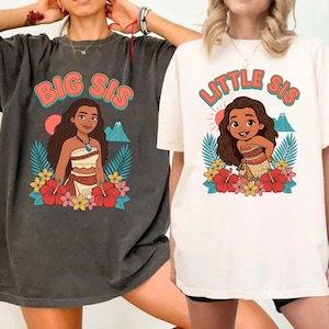 May include: Two t-shirts, one dark gray and one off-white, featuring a cartoon illustration of a girl surrounded by flowers and tropical leaves. The dark gray shirt says "BIG SIS" and the off-white shirt says "LITTLE SIS" in a colorful font.