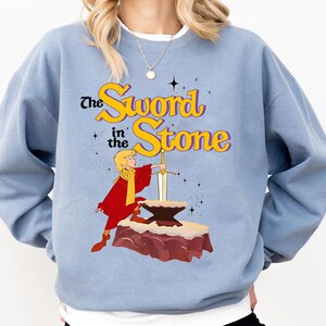 K&ouml;nnte beinhalten: Hellblaues Sweatshirt mit dem Text "The Sword in the Stone" in Gelb und Lila. Die Grafik zeigt eine Zeichentrickfigur in Rot, die ein Schwert in einem Stein h&auml;lt. Das Sweatshirt hat lange &Auml;rmel.