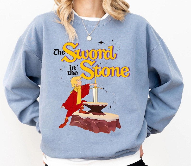 K&ouml;nnte beinhalten: Hellblaues Sweatshirt mit dem Text "The Sword in the Stone" in Gelb und Lila. Die Grafik zeigt eine Zeichentrickfigur in Rot, die ein Schwert in einem Stein h&auml;lt. Das Sweatshirt hat lange &Auml;rmel.