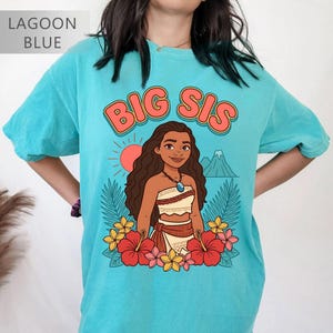 May include: Lagoon blue t-shirt with the words "BIG SIS" in orange lettering. The shirt features a cartoon illustration of a woman with long brown hair, a necklace, and a floral skirt. Red and yellow flowers surround the illustration.