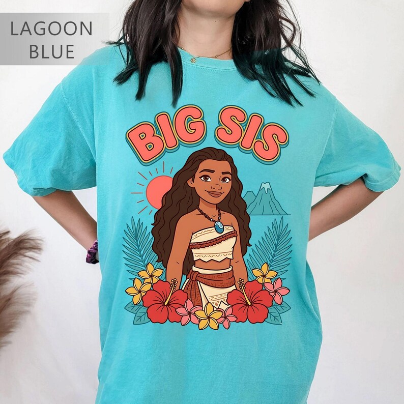 May include: Lagoon blue t-shirt with the words "BIG SIS" in orange lettering. The shirt features a cartoon illustration of a woman with long brown hair, a necklace, and a floral skirt. Red and yellow flowers surround the illustration.