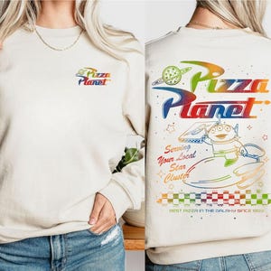 Puede incluir: Una sudadera blanca con un gr&aacute;fico de arco&iacute;ris del texto "Pizza Planet" y un personaje de dibujos animados sosteniendo una caja de pizza. El texto "Serving Your Local Star Cluster" y "Best Pizza in the Galaxy Since 1995" tambi&eacute;n est&aacute;n incluidos en el gr&aacute;fico.