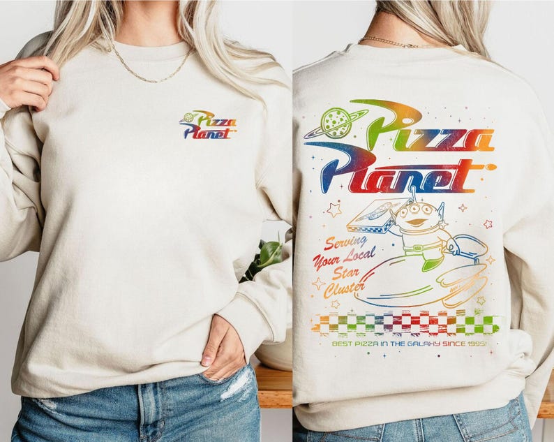 Puede incluir: Una sudadera blanca con un gr&aacute;fico de arco&iacute;ris del texto "Pizza Planet" y un personaje de dibujos animados sosteniendo una caja de pizza. El texto "Serving Your Local Star Cluster" y "Best Pizza in the Galaxy Since 1995" tambi&eacute;n est&aacute;n incluidos en el gr&aacute;fico.