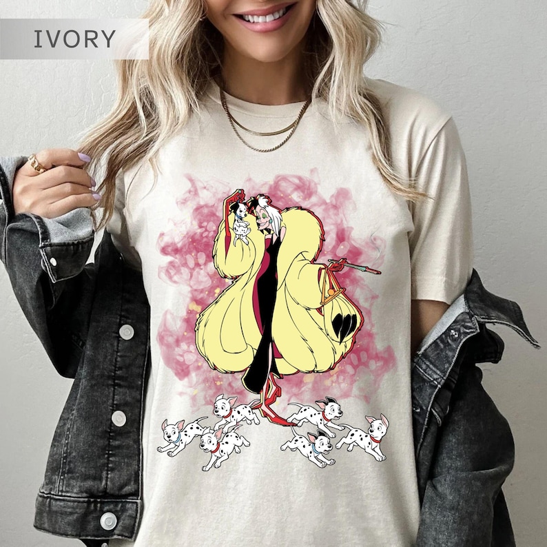 K&ouml;nnte beinhalten: Elfenbeinfarbenes T-Shirt mit einer Cartoon-Illustration einer Figur in einem gelben Mantel und schwarzen Kleid, umgeben von Dalmatiner-Welpen. Das Design ist vor einem rosa und wei&szlig;en Hintergrund angeordnet.