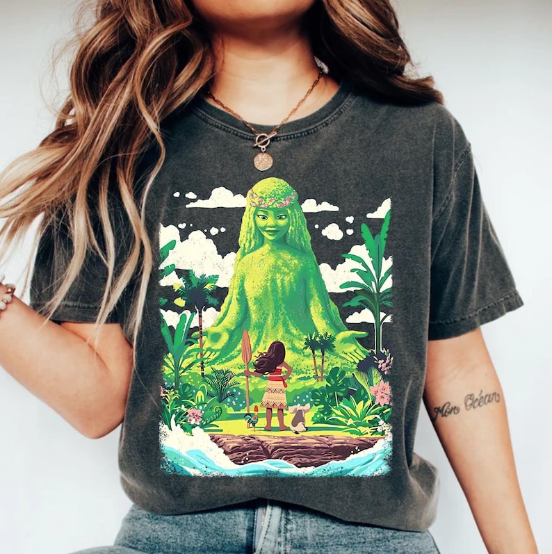 Puede incluir: Una camiseta gris oscuro con un estampado gr&aacute;fico de una figura de diosa verde con una corona de flores. La diosa est&aacute; sentada en una postura de yoga con una figura m&aacute;s peque&ntilde;a de una mujer de pie frente a ella. El gr&aacute;fico est&aacute; rodeado de plantas y flores tropicales. La camiseta tiene un aspecto desgastado.