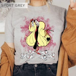 K&ouml;nnte beinhalten: Sportgraues T-Shirt mit einer Cartoon-Illustration einer Figur in einem gelben Mantel, einem schwarzen Kleid und roten Schuhen, die einen kleinen Hund und eine Tasche h&auml;lt, umgeben von laufenden Dalmatiner-Welpen. Der Text "SPORT GREY" steht oben.