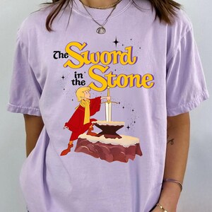 K&ouml;nnte beinhalten: Ein helllila T-Shirt mit einer Grafik aus "The Sword in the Stone". Das Design zeigt eine Figur in roter und gelber Kleidung, die ein Schwert aus einem Amboss zieht. Der Text "The Sword in the Stone" ist gelb und schwarz.