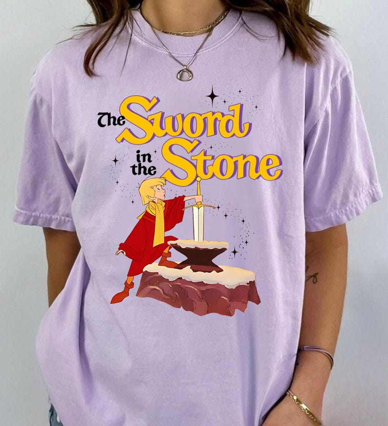 K&ouml;nnte beinhalten: Ein helllila T-Shirt mit einer Grafik aus "The Sword in the Stone". Das Design zeigt eine Figur in roter und gelber Kleidung, die ein Schwert aus einem Amboss zieht. Der Text "The Sword in the Stone" ist gelb und schwarz.