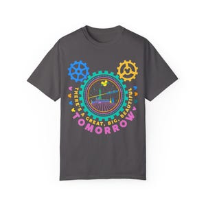 May include: A dark grey short-sleeve t-shirt featuring a colourful graphic of gears and a futuristic building. The text reads, "THERE'S A GREAT, BIG, BEAUTIFUL TOMORROW" in a circular arrangement around the central illustration with small mouse icons.