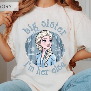 May include: Ivory-colored t-shirt featuring a cartoon illustration of Elsa from Frozen. The design includes the text "big sister, I'm her elsa" in light blue, with snowflakes and a winter scene background. The shirt is a casual wear item.