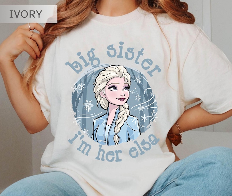 May include: Ivory-colored t-shirt featuring a cartoon illustration of Elsa from Frozen. The design includes the text "big sister, I'm her elsa" in light blue, with snowflakes and a winter scene background. The shirt is a casual wear item.