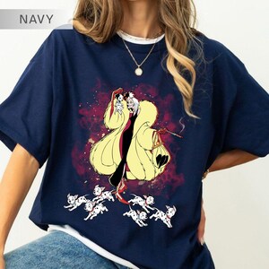 K&ouml;nnte beinhalten: Marineblaues T-Shirt mit einer Cartoon-Illustration einer Figur in einem gelben Mantel, die eine Zigarette und eine Tasche h&auml;lt, umgeben von Dalmatiner-Welpen. Das T-Shirt hat einen wei&szlig;en Kragen und eine goldene Halskette.