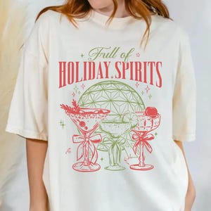 May include: An off-white short-sleeve t-shirt featuring a graphic print with the text "Full of HOLIDAY SPIRITS" in red and green. The design includes a green disco ball and three cocktail glasses decorated with red bows and garnishes.