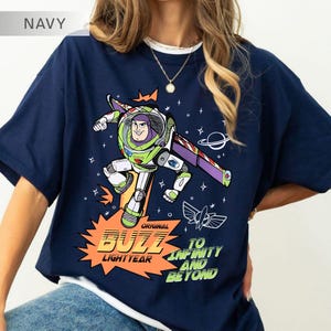 K&ouml;nnte beinhalten: Marineblaues T-Shirt mit einem Aufdruck von Buzz Lightyear aus Toy Story. Das Design zeigt Buzz in seinem Raumanzug, die Worte "Original Buzz Lightyear" und "To Infinity and Beyond". Das Shirt hat einen Rundhalsausschnitt und kurze &Auml;rmel.