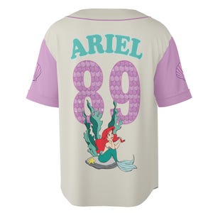 May include: Cream-colored baseball jersey with light purple sleeves and collar. The back features the name "ARIEL" in teal, the number "89" in a mermaid scale pattern, and a graphic of Ariel the Little Mermaid.