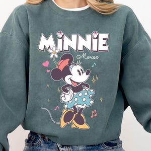 Puede incluir: Una sudadera azul verdosa con un estampado gr&aacute;fico de Minnie Mouse. El gr&aacute;fico presenta a Minnie Mouse con un vestido de lunares y una flor en el pelo. El texto "Minnie Mouse" est&aacute; impreso encima de ella.