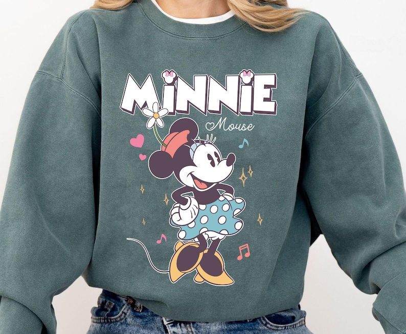 Puede incluir: Una sudadera azul verdosa con un estampado gr&aacute;fico de Minnie Mouse. El gr&aacute;fico presenta a Minnie Mouse con un vestido de lunares y una flor en el pelo. El texto "Minnie Mouse" est&aacute; impreso encima de ella.