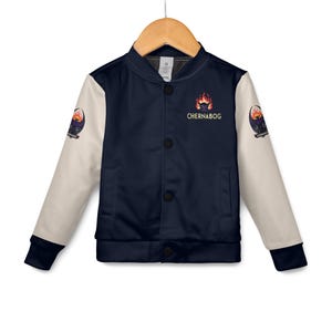 May include: A navy blue and cream-colored bomber jacket with black buttons. The jacket features the word "Chernabog" above a fiery graphic on the chest. The sleeves have a patch with a similar design. The jacket is hanging on a wooden hanger.