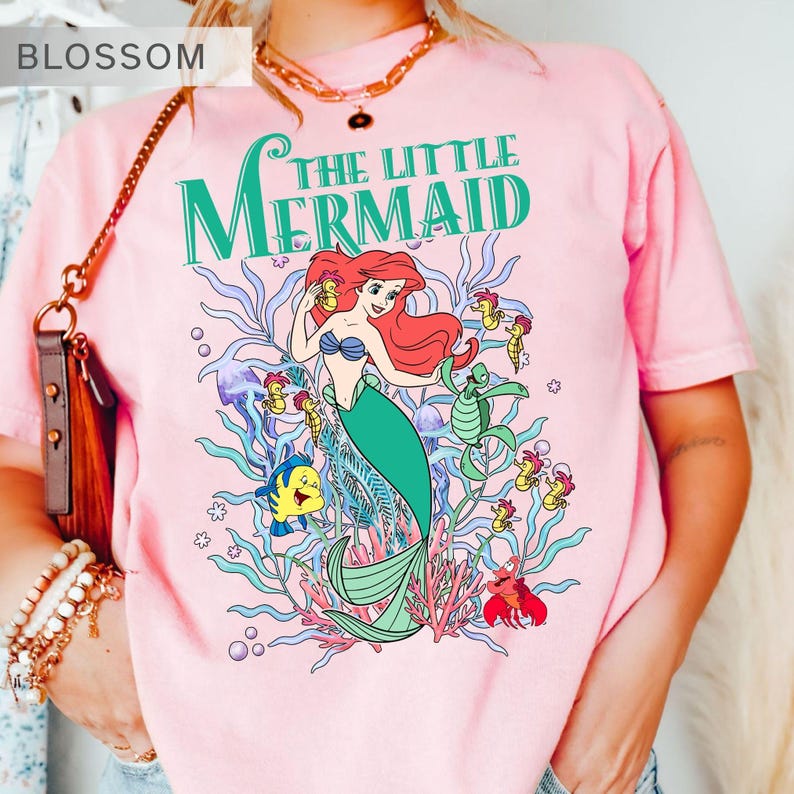 May include: Pink t-shirt featuring a colorful graphic of Ariel from The Little Mermaid. The design includes the text "The Little Mermaid" in green, along with illustrations of sea creatures like Flounder, seahorses, and a crab.