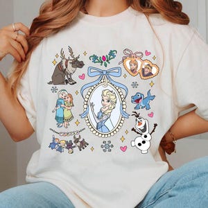 Puede incluir: Camiseta color crema con ilustraciones de dibujos animados de personajes de la pel&iacute;cula animada "Frozen". El dise&ntilde;o incluye im&aacute;genes de Elsa, Anna, Olaf, Sven y otros elementos, con un lazo azul y detalles decorativos.