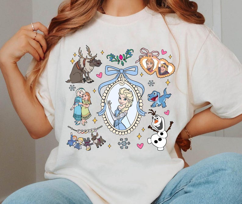 Puede incluir: Camiseta color crema con ilustraciones de dibujos animados de personajes de la pel&iacute;cula animada "Frozen". El dise&ntilde;o incluye im&aacute;genes de Elsa, Anna, Olaf, Sven y otros elementos, con un lazo azul y detalles decorativos.