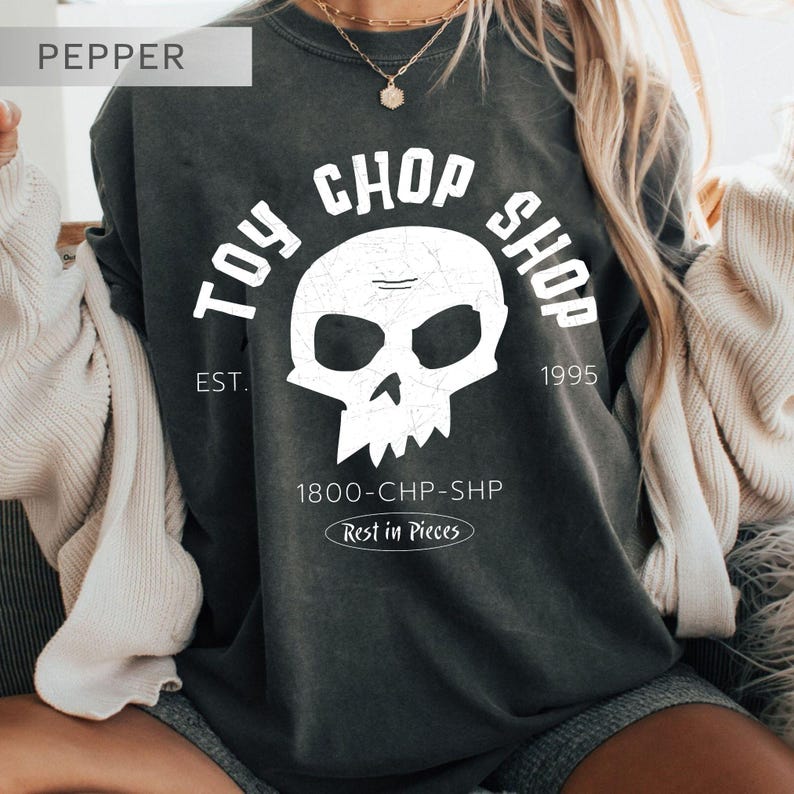 Pu&ograve; includere: Una t-shirt girocollo grigio scuro con una grafica bianca raffigurante un teschio. Il testo recita TOY CHOP SHOP, EST. 1995, 1800-CHP-SHP e Rest in Pieces. Il design presenta una texture usurata. La maglia &egrave; indossata con un cardigan color crema.