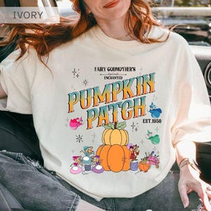 Puede incluir: Sudadera de color marfil con un gráfico colorido que dice "Fairy Godmother's Enchanted Pumpkin Patch Est. 1950". El diseño presenta una calabaza, personajes de hadas y estrellas. La camisa es una parte superior informal de manga larga.