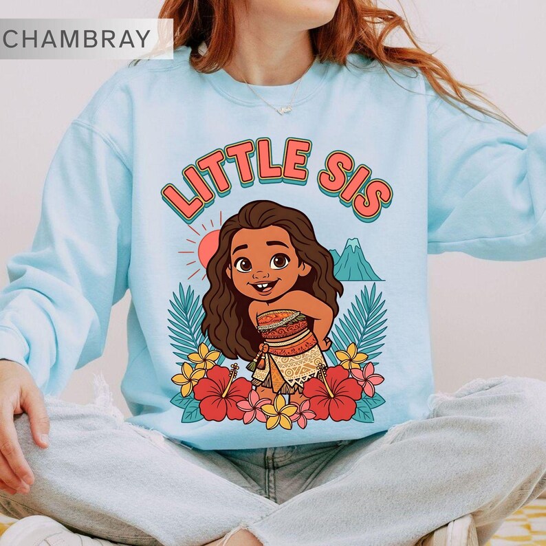 May include: Light blue sweatshirt with the text "LITTLE SIS" in orange and a cartoon illustration of a girl surrounded by flowers and tropical leaves. The design includes a sun and a mountain.