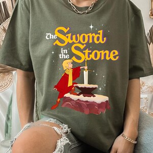 K&ouml;nnte beinhalten: Olivgr&uuml;nes T-Shirt mit einer Grafik aus "The Sword in the Stone". Das Design zeigt eine Zeichentrickfigur in roter und gelber Kleidung, die ein Schwert aus einem Stein zieht. Der Text "The Sword in the Stone" ist in Gelb und Lila gehalten.