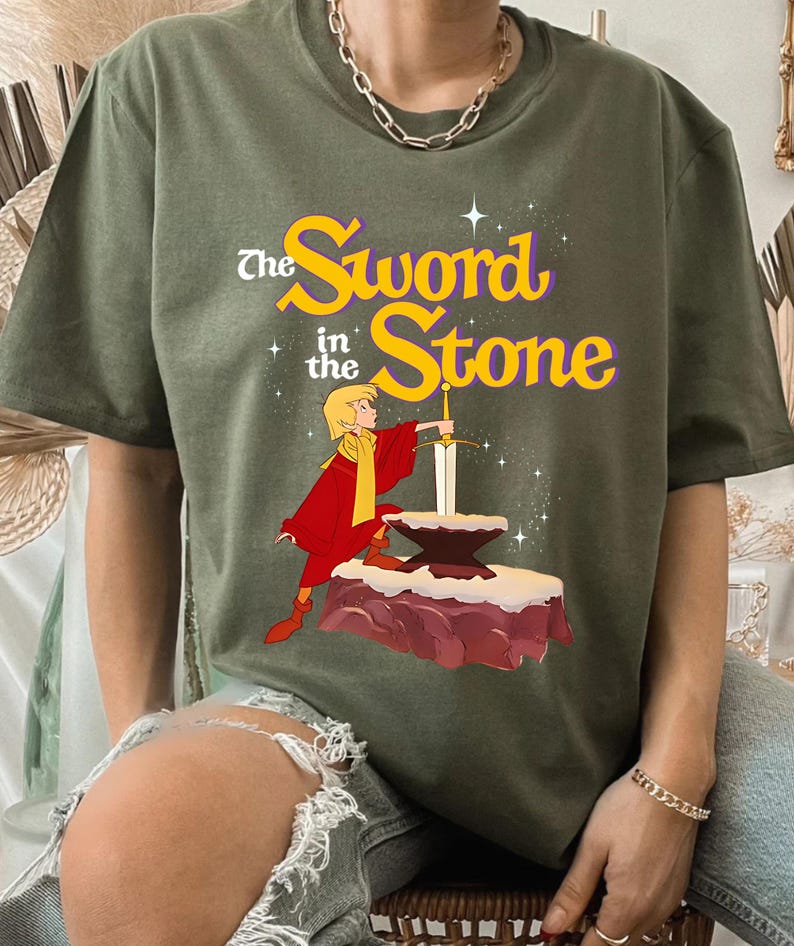 K&ouml;nnte beinhalten: Olivgr&uuml;nes T-Shirt mit einer Grafik aus "The Sword in the Stone". Das Design zeigt eine Zeichentrickfigur in roter und gelber Kleidung, die ein Schwert aus einem Stein zieht. Der Text "The Sword in the Stone" ist in Gelb und Lila gehalten.