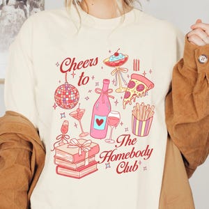 May include: Cream-colored t-shirt with a graphic design featuring a disco ball, wine bottle, pizza slice, and other party-themed illustrations. The text "Cheers to The Homebody Club" is also displayed.