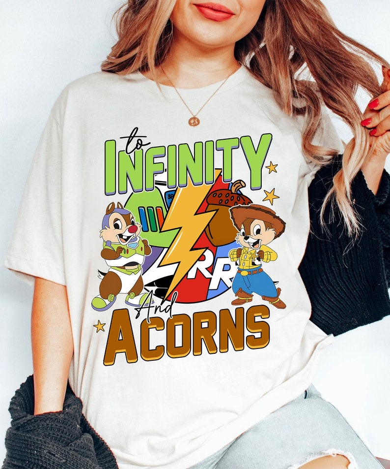 May include: White t-shirt with a graphic design featuring two cartoon chipmunks, one dressed as Buzz Lightyear and the other as Woody from Toy Story. The design also includes a lightning bolt and the text "to infinity and acorns".