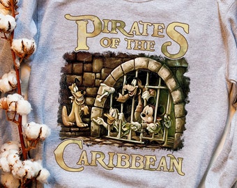 Könnte beinhalten: Ein graues Sweatshirt mit einem grafischen Aufdruck von Mickey Mouse, Donald Duck und Goofy, die als Piraten verkleidet sind. Der Text "Pirates of the Caribbean" ist in goldenen Buchstaben gedruckt.