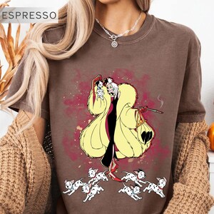 K&ouml;nnte beinhalten: Espressofarbenes T-Shirt mit einer Cartoon-Illustration einer Figur in einem gelben Mantel, einem schwarzen Kleid und roten Schuhen, die einen kleinen Hund und eine Zigarette h&auml;lt. Mehrere Dalmatinerwelpen befinden sich zu F&uuml;&szlig;en der Figur. Das Wort "ESPRESSO" steht oben links.