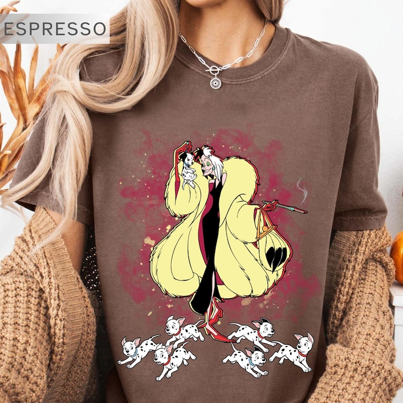 K&ouml;nnte beinhalten: Espressofarbenes T-Shirt mit einer Cartoon-Illustration einer Figur in einem gelben Mantel, einem schwarzen Kleid und roten Schuhen, die einen kleinen Hund und eine Zigarette h&auml;lt. Mehrere Dalmatinerwelpen befinden sich zu F&uuml;&szlig;en der Figur. Das Wort "ESPRESSO" steht oben links.